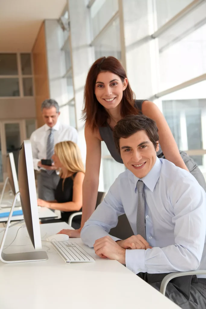 Two office workers in the background looking at a computer screen and talking to each other. In the foreground, a man and a woman are looking at the camera and smiling. The man is sitting at a desk using a computer and the woman is standing behind him.